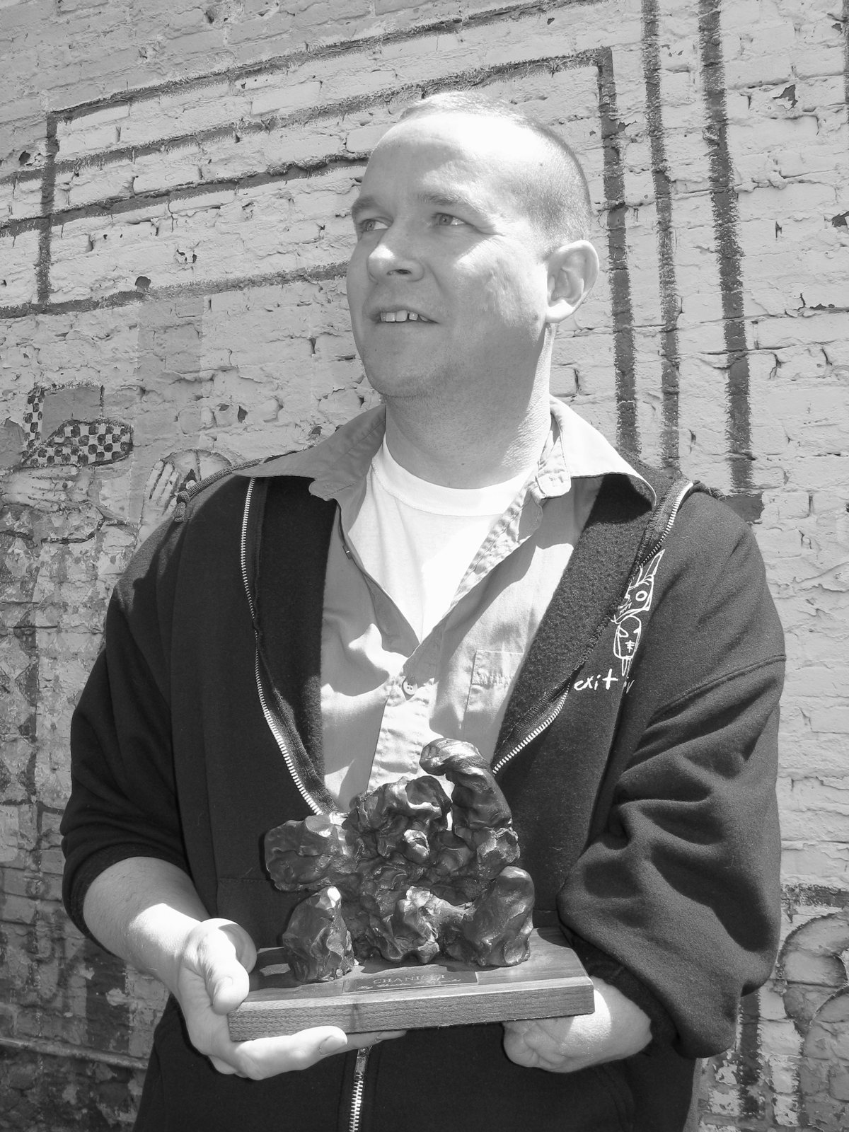 Black and white photo of Matt Sesow holding the CHANISKI bronze in front of a painted brick wall